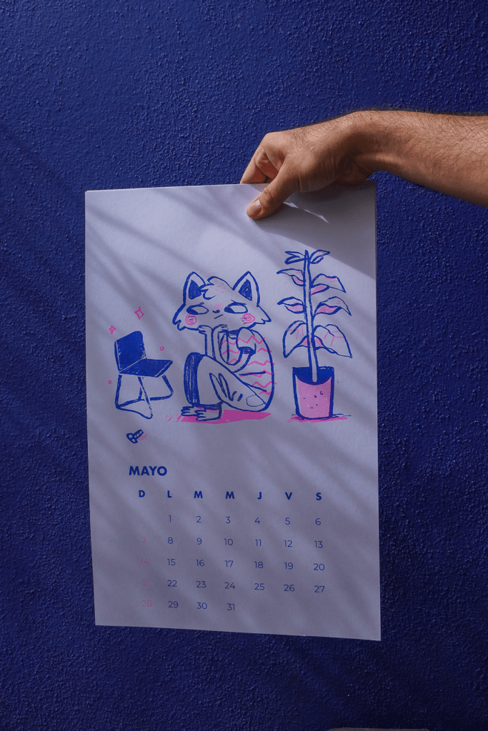 A hand holding a printed calendar for May, featuring an illustration of a cat-like character sitting next to a plant, against a blue wall.