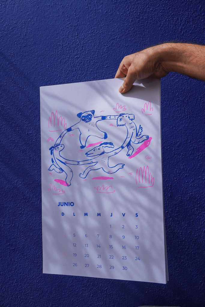 A person holding a colorful June calendar featuring a playful illustration of two animals interacting, with a blue background.