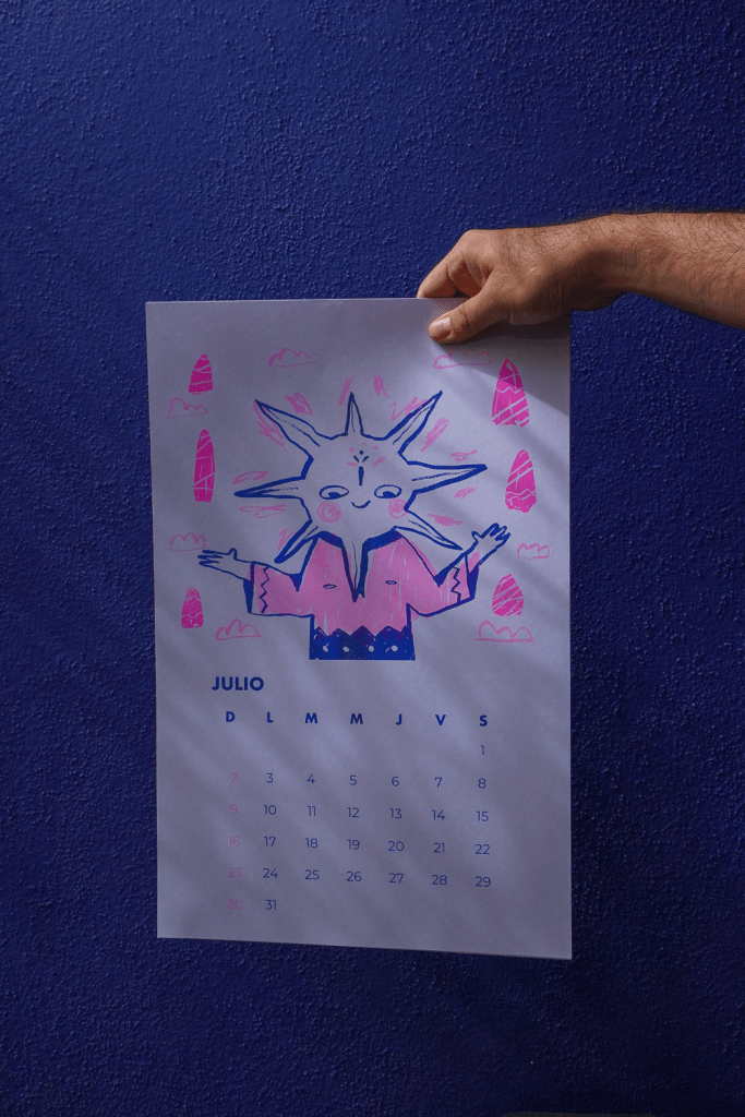 A hand holding a colorful calendar page for July featuring a cartoon sun with arms, surrounded by clouds and geometric shapes, against a blue wall.