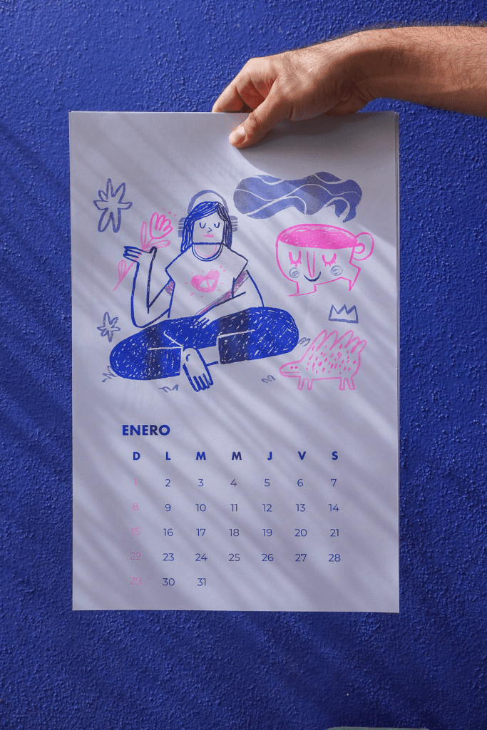 A hand holding a colorful illustrated calendar for January, featuring a person meditating with headphones, surrounded by playful doodles and a cup.