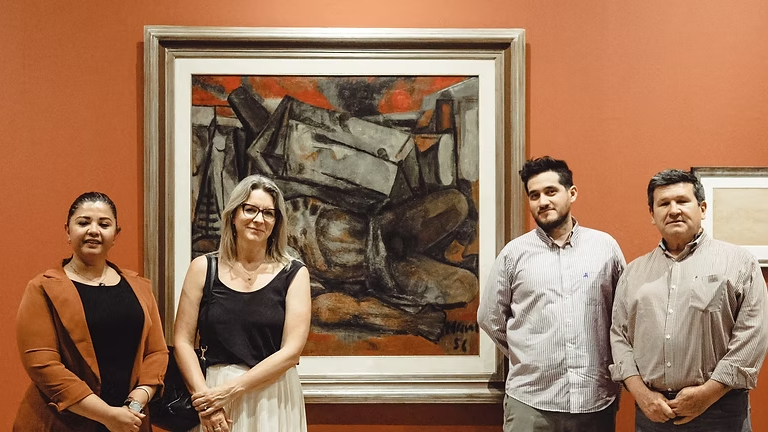 Cuatro personas posando en una galería de arte, frente a una pintura enmarcada de un artista, con paredes de color rojo.