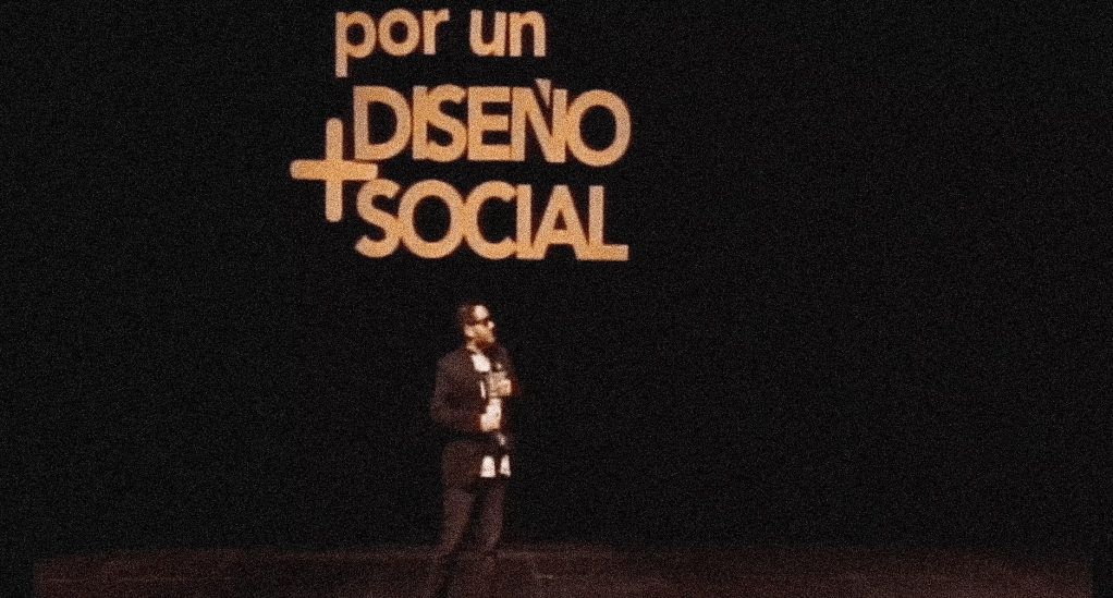 Un hombre de pie en un escenario frente a una gran pantalla con el texto 'por un Diseño Social' en letras amarillas sobre un fondo oscuro.