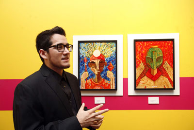 A man wearing glasses and a black suit stands beside two colorful paintings on a vibrant yellow wall, gesturing as he talks.