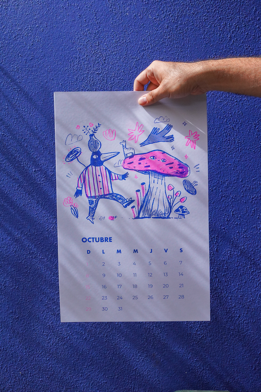 Calendario de octubre con ilustraciones coloridas de un personaje y un hongo, sostenido por una mano frente a una pared azul.