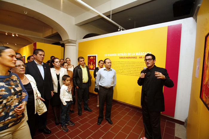 A gallery event with a speaker addressing an audience in front of a colorful exhibition wall, featuring a display titled '15 tesoros detrás de la máscara' by Carlos Hernández.