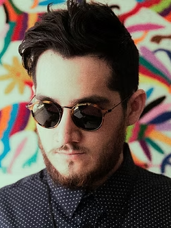 Retrato de un hombre con barba y lentes de sol frente a un bordado de colores